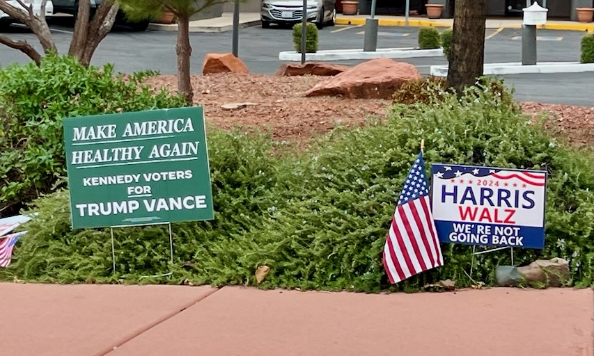 A green "Make America Healthy Again" Kennedy Voters for Trump Vance sign competes with a Harris Walz "We're not going back" sign.