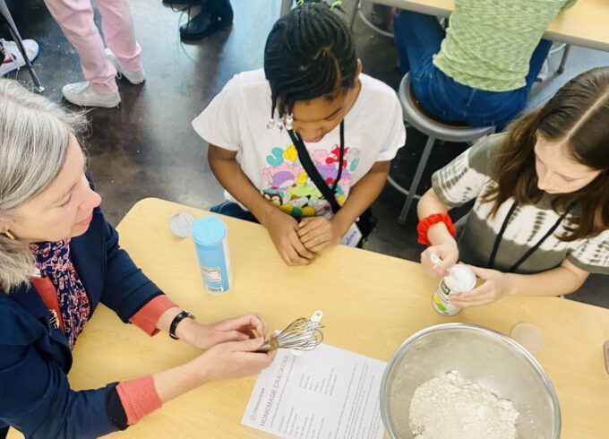 USDA's Stacy Dean sits with two fifth graders at a table - they are mixing the ingredients for whole wheat crackers in a stainless steal bowl.
