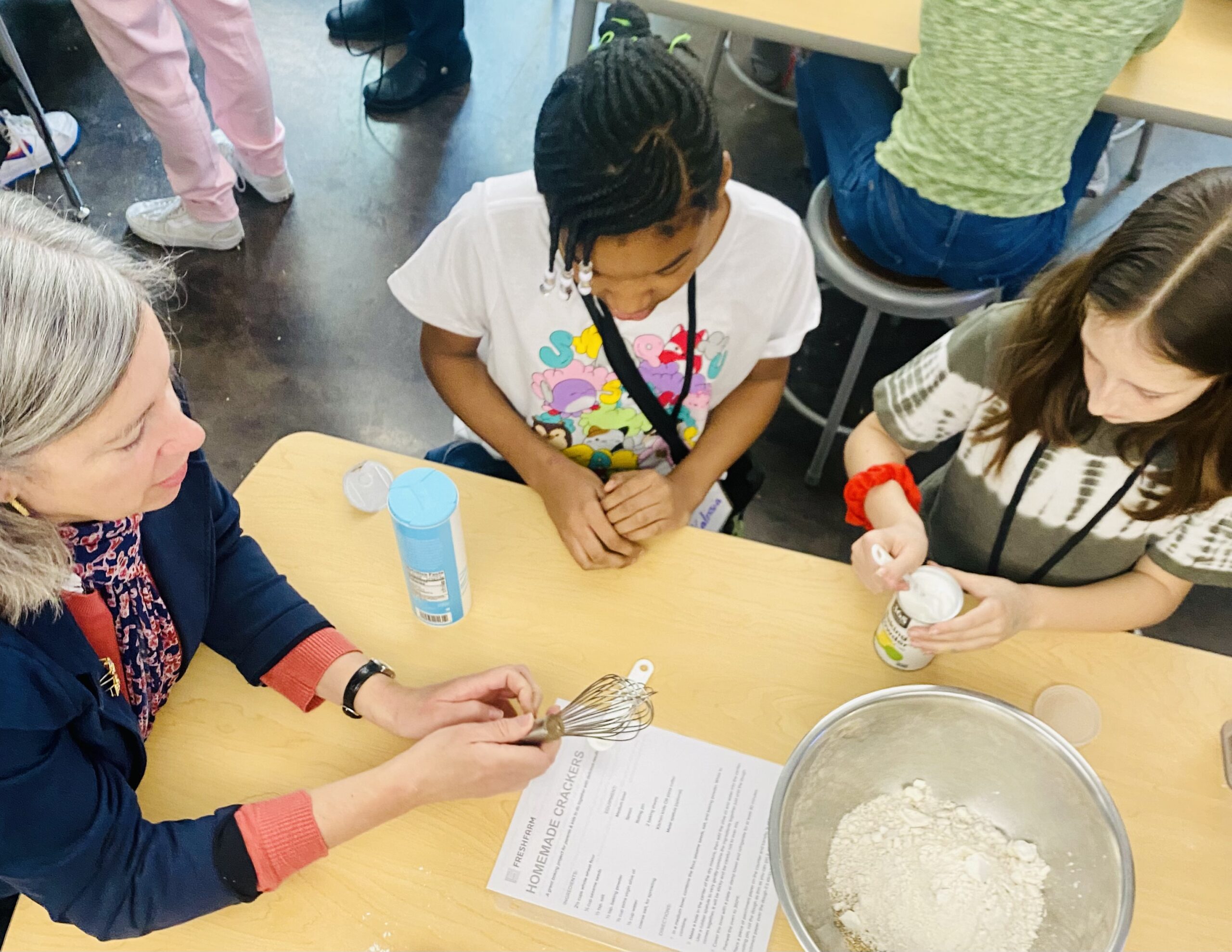 USDA's Stacy Dean sits with two fifth graders at a table - they are mixing the ingredients for whole wheat crackers in a stainless steal bowl.