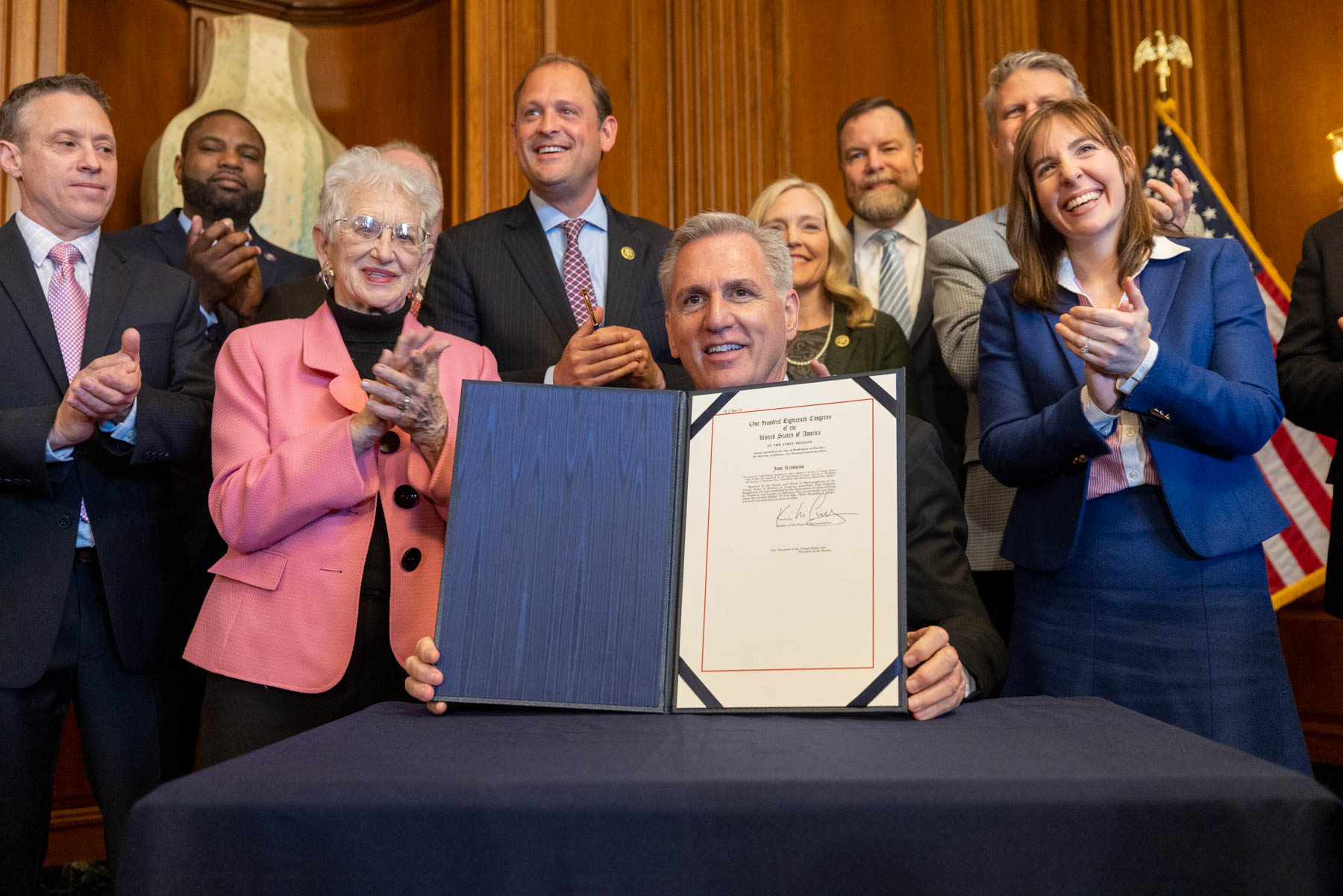 A photo of House Speaker Kevin McCarthy holding up a signed bill at a table surrounded by lawmakers.