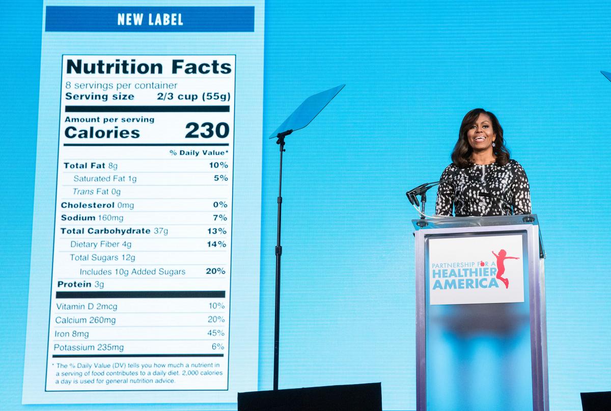 A photo of former first lady Michelle Obama speaking on stage with a large display of the Nutrition Facts label to her right.