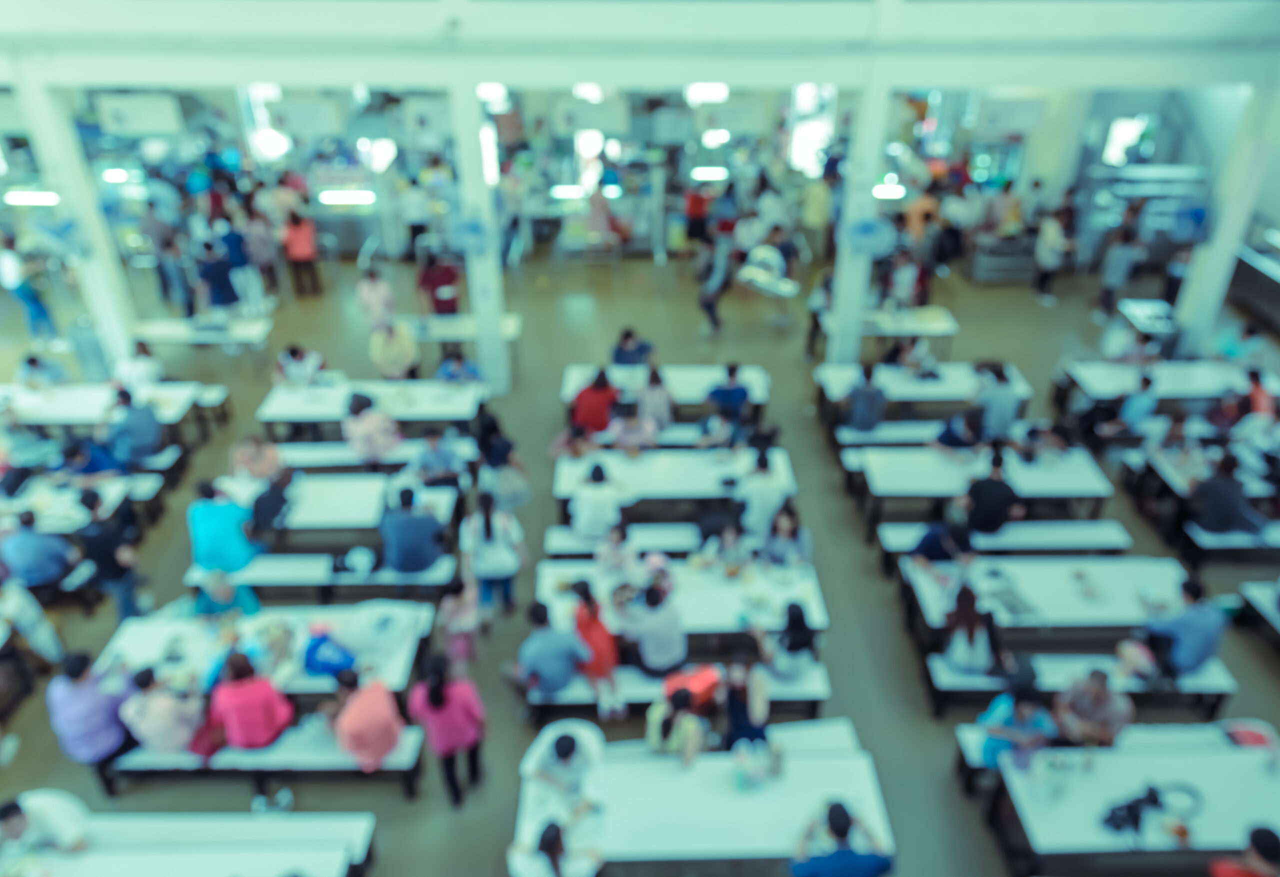 A blurred aerial photo of a school cafeteria, in which kids are eating at tables.