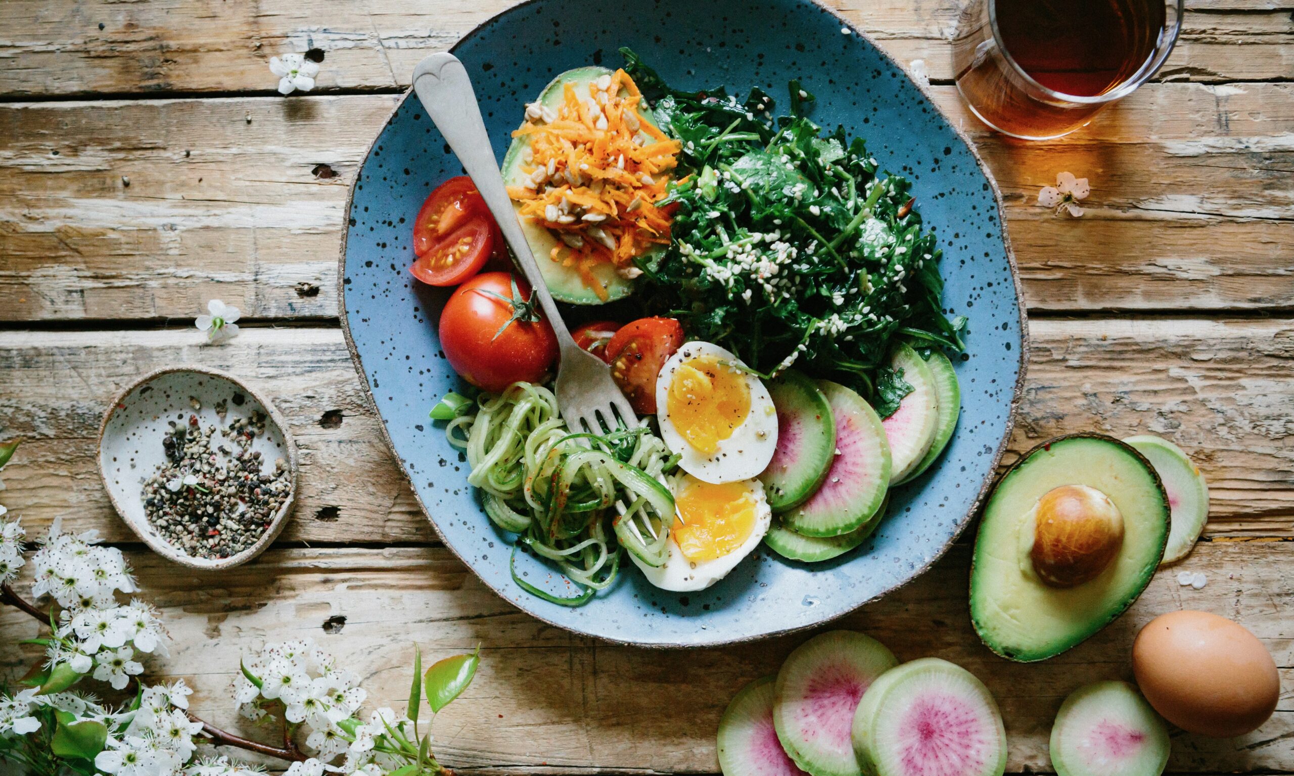 A blue ceramic bowl sitting on a wood table is filled with an avocado, an egg, radishes, sauteed greens and tomatoes. An avocado and radishes are featured on the table next to the bowl.