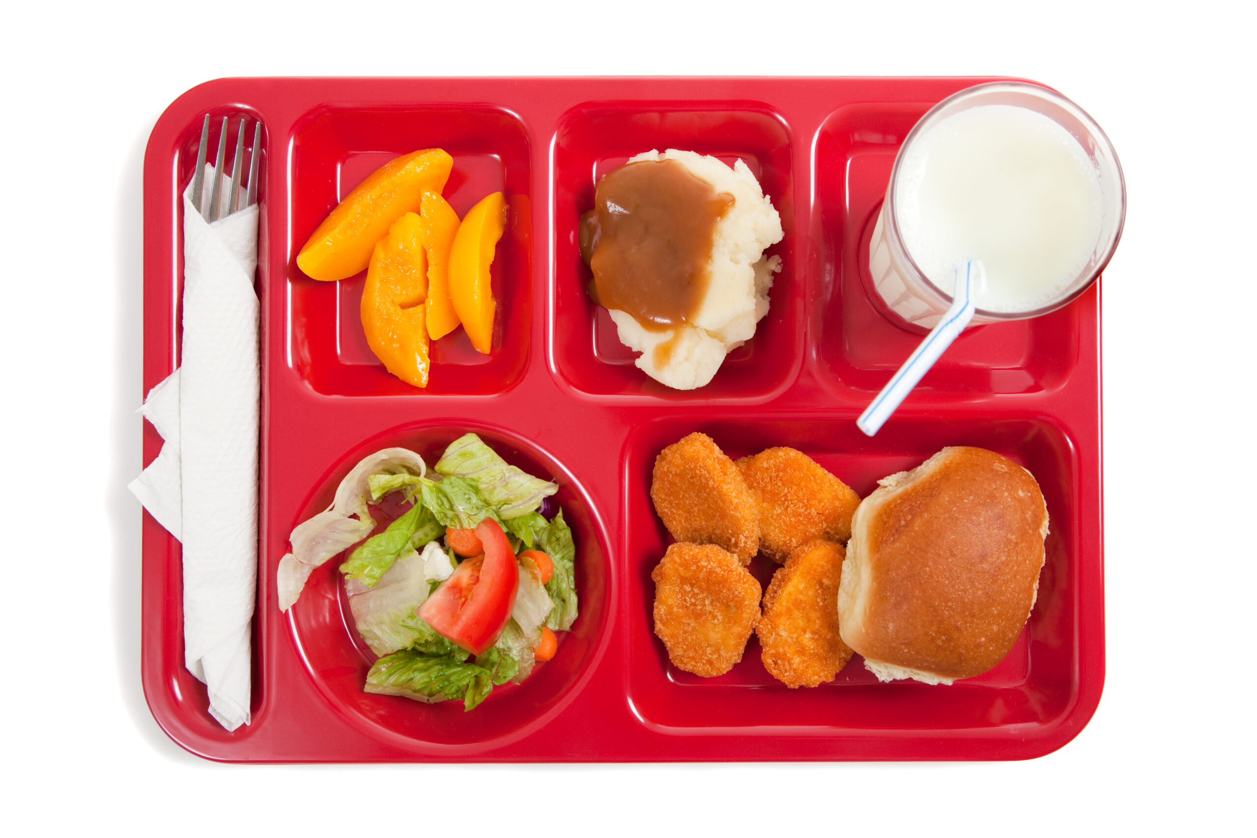 A red school lunch tray viewed from above. The tray contains a roll, chicken nuggets, salad, peaches, mashed potatoes and milk.